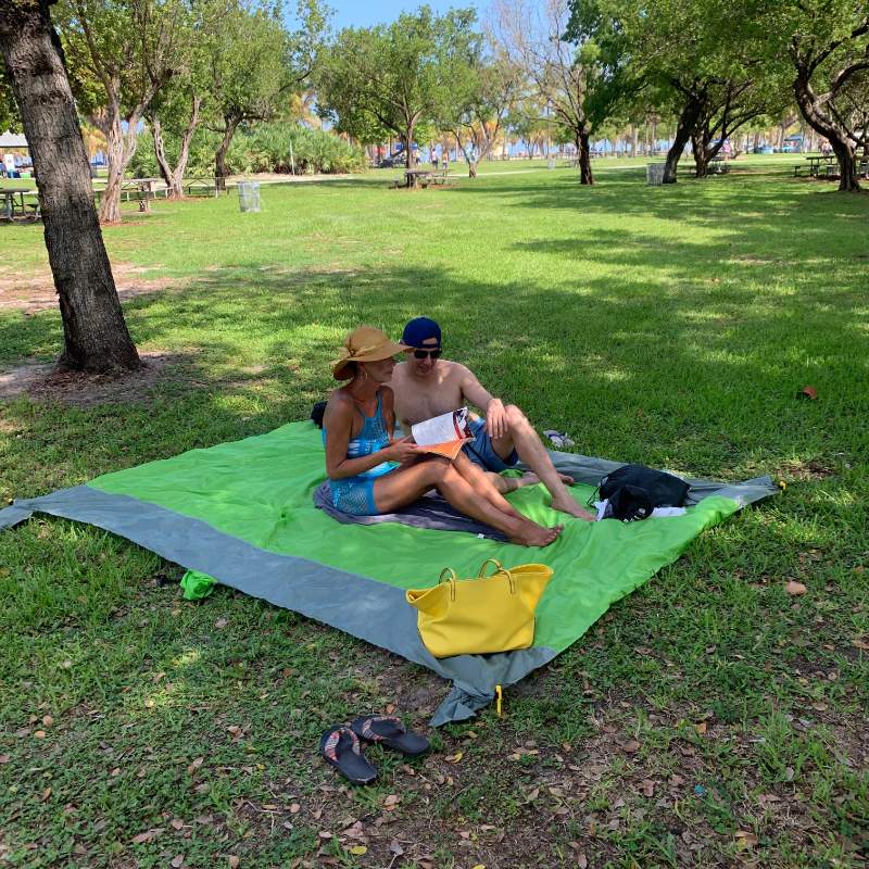 Two people reading on a light green SandStop blanket under trees